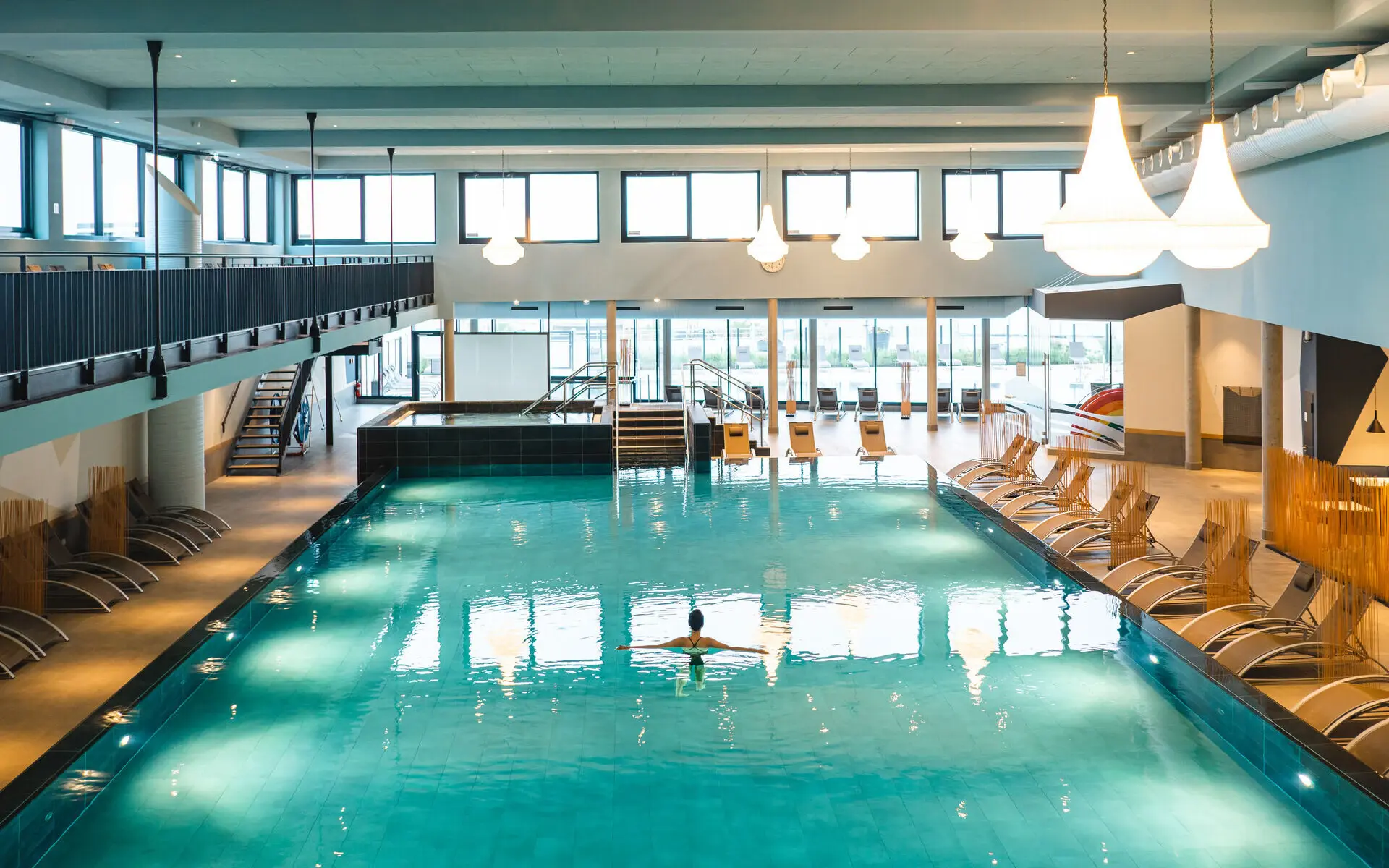 Innenpool aja Travemünde Eine Frau steht mit ausgebreiteten Armen im weitläufigen Innenpool des aja Travemünde. Deckenleuchten und Randbeleuchtung des Pools erhellen den Raum. Am Becken rand stehen einige Poolliegen aufgereiht, welche jedoch nicht besetztt sind. Im Hintergrund ist der Zugang über eine kleine Treppe zum Whirlpool sowie das Panoramafenster mit Blick auf den Außenbereich zu sehen.
