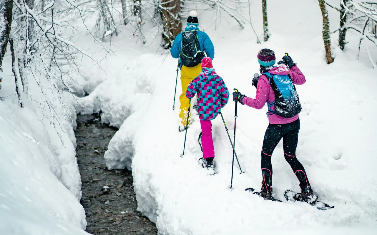 Drei Personen stapfen mit Schneeschuhen einen kleinen Bach durch den Schnee entlang.