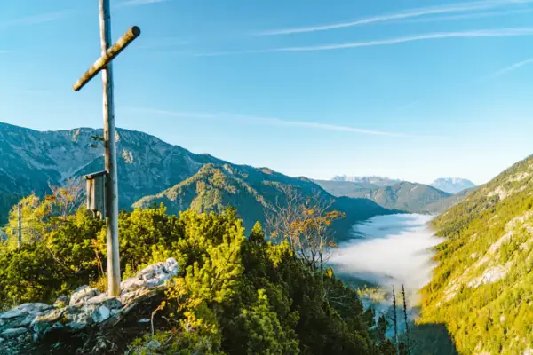 Gipfelkreuz Blick auf ein Tal mit Bäumen und Bergen im Hintergrund.