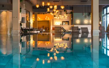 Innenpool aja Garmisch-Partenkirchen mit umstehenden Liegen und warm leuchtenden Deckenlampen, die sich auf der Wasseroberfläche spiegeln.