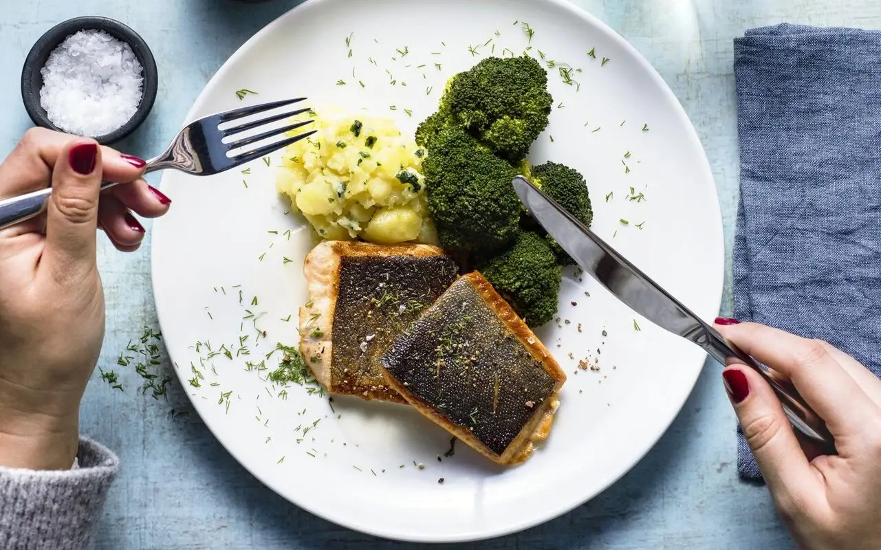 Abendessen aja Grömitz Eine Frau sitzt an einem Tisch vor einem Teller mit Fischfilet, Brokkoli und Kartoffelpüree und hält Besteck in ihren Händen.
