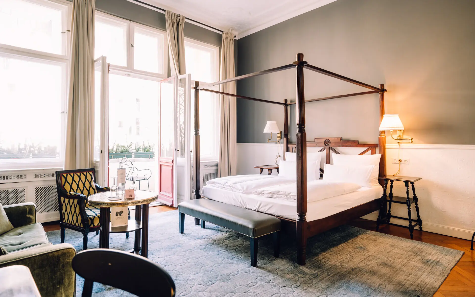 HENRI Berlin Zimmer Elegantes Hotelzimmer im HENRI Berlin mit einem stilvollen Himmelbett, klassischen Holzmöbeln, einem gemütlichen Sessel, einem kleinen Tisch und großen Fenstern, die viel Tageslicht einlassen, sowie einem Zugang zu einem Balkon.
