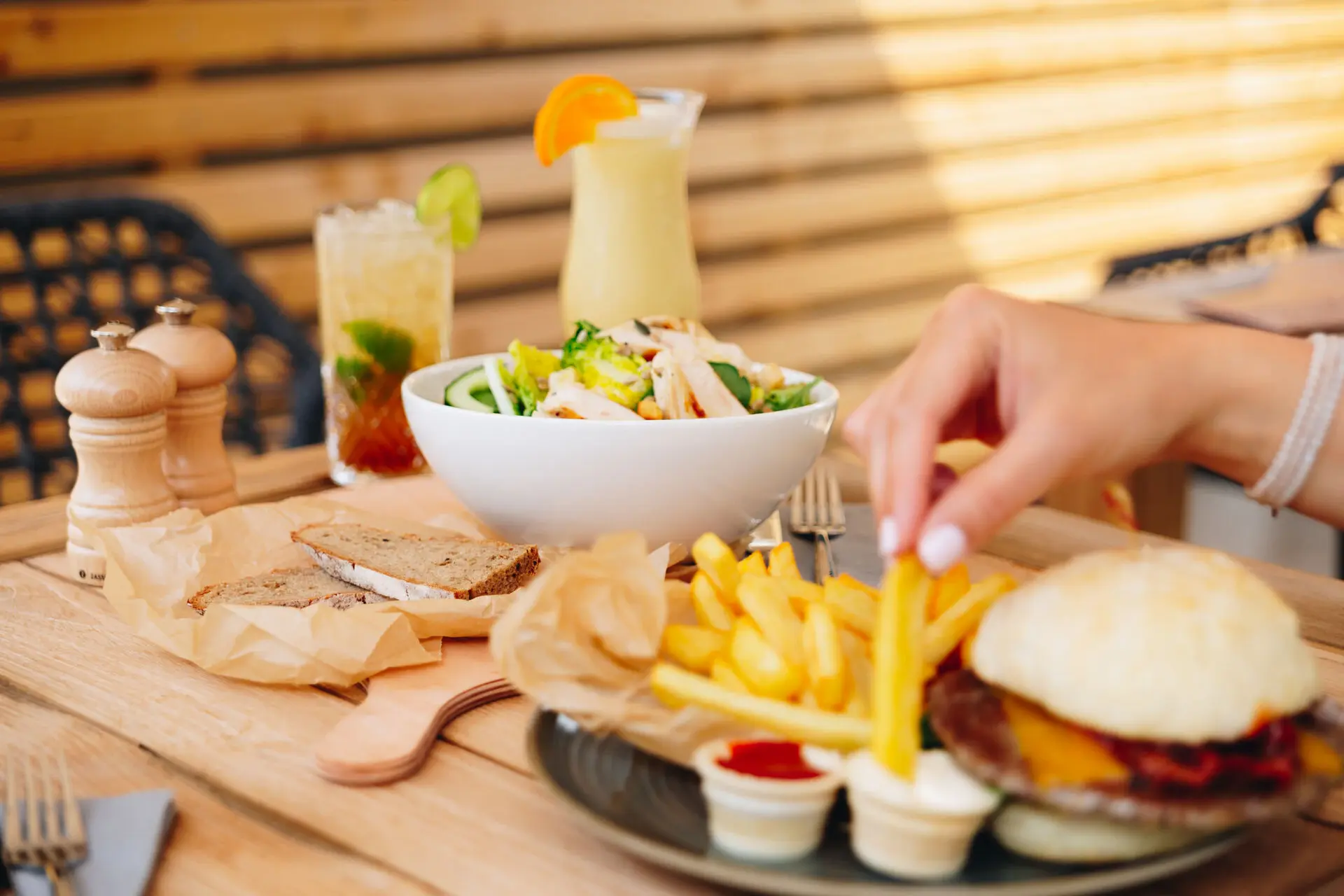 Ein gedeckter Holztisch mit einer Auswahl an Speisen: ein knackiger Salat mit Hähnchenstreifen in einer weißen Schüssel, ein Burger mit Pommes frites und verschiedenen Dips, begleitet von frischem Brot. Im Hintergrund stehen zwei erfrischende Cocktails mit Garnitur.