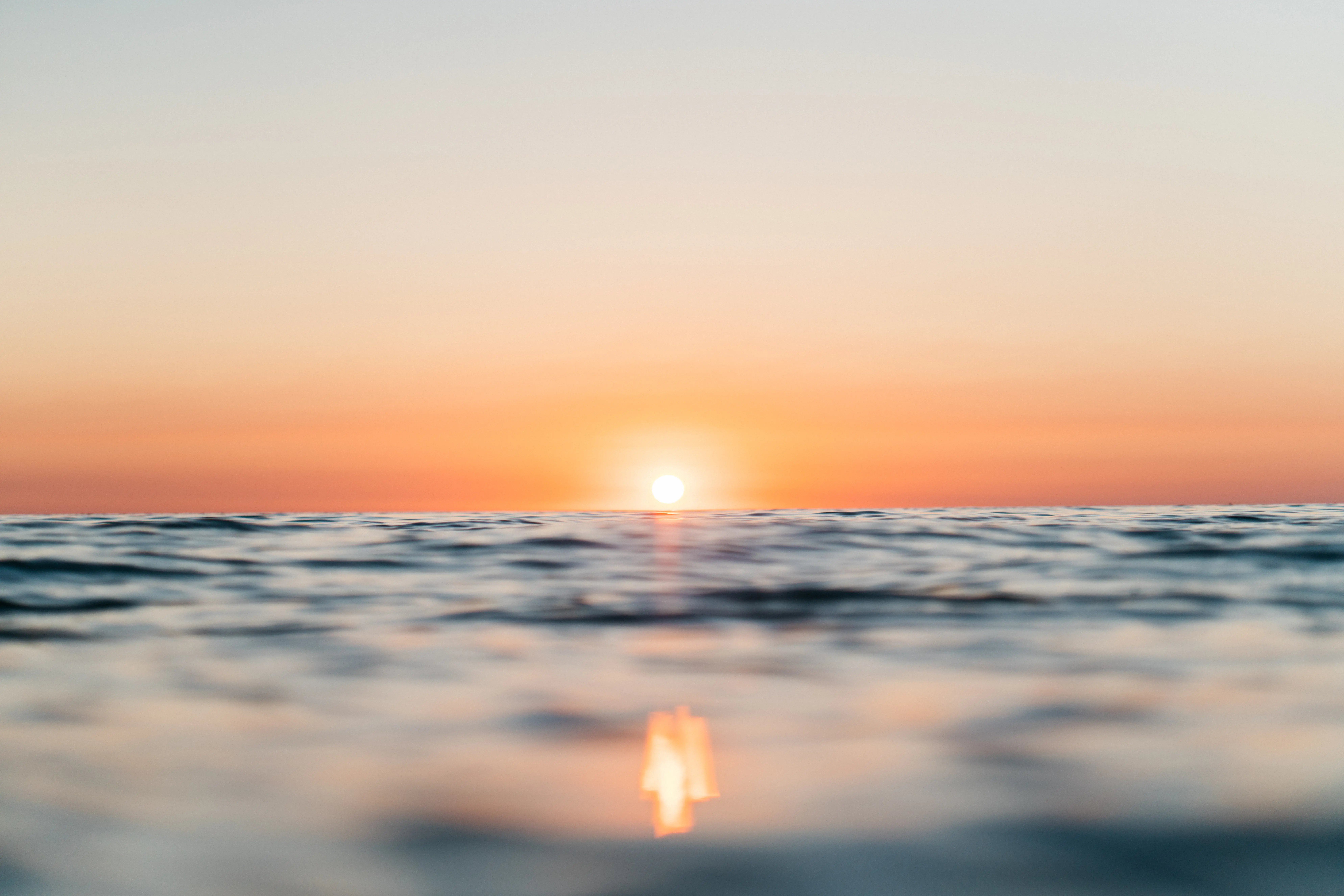 Sonnenuntergang über dem Meer, die Sonne kurz über dem Horizont, das Wasser im Vordergrund sanft bewegt, unter einem pastellfarbenen Himmel.
