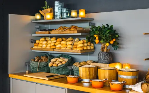 Ein warm beleuchteter Eckbereich eine Buffets mit Regalen, auf denen sich Brötchen und Gebäcksorten befinden. Daneben steht ein kleiner Orangenbaum. Ein warm beleuchteter Eckbereich eine Buffets mit Regalen, auf denen sich Brötchen und Gebäcksorten befinden. Daneben steht ein kleiner Orangenbaum.