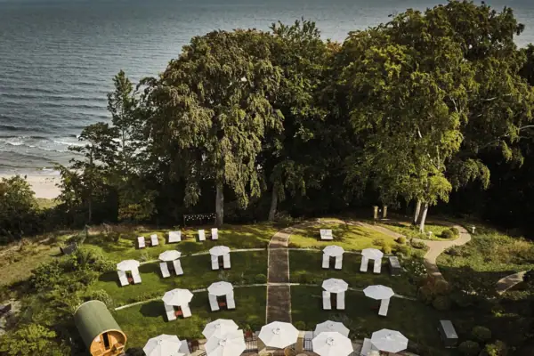 Garten Nordperd & Villen Eine Gruppe weißer Sonnenschirme auf einer grasbewachsenen Fläche mit Bäumen und dem Meer im Hintergrund.
