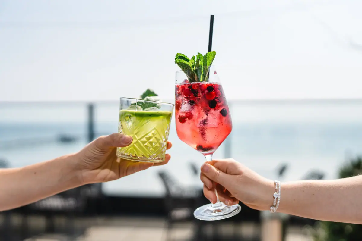 Drinks Rooftop Bar Grömitz Ein Paar Hände hält Cocktails mit Früchtegarnitur auf einer Rooftop Bar mit Meerblick.