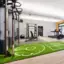 In einem hellen Raum mit Holzboden und weißen Wänden stehen mehrere Cardio- und Kraftgeräte aufgereiht. Die rechte Wand mit zwei großen Fenstern ist orange gestrichen und im Vordergrund liegt ein grasgrüner Teppich mit einigen weißen Markierungen vor einem Fitnessgerät.