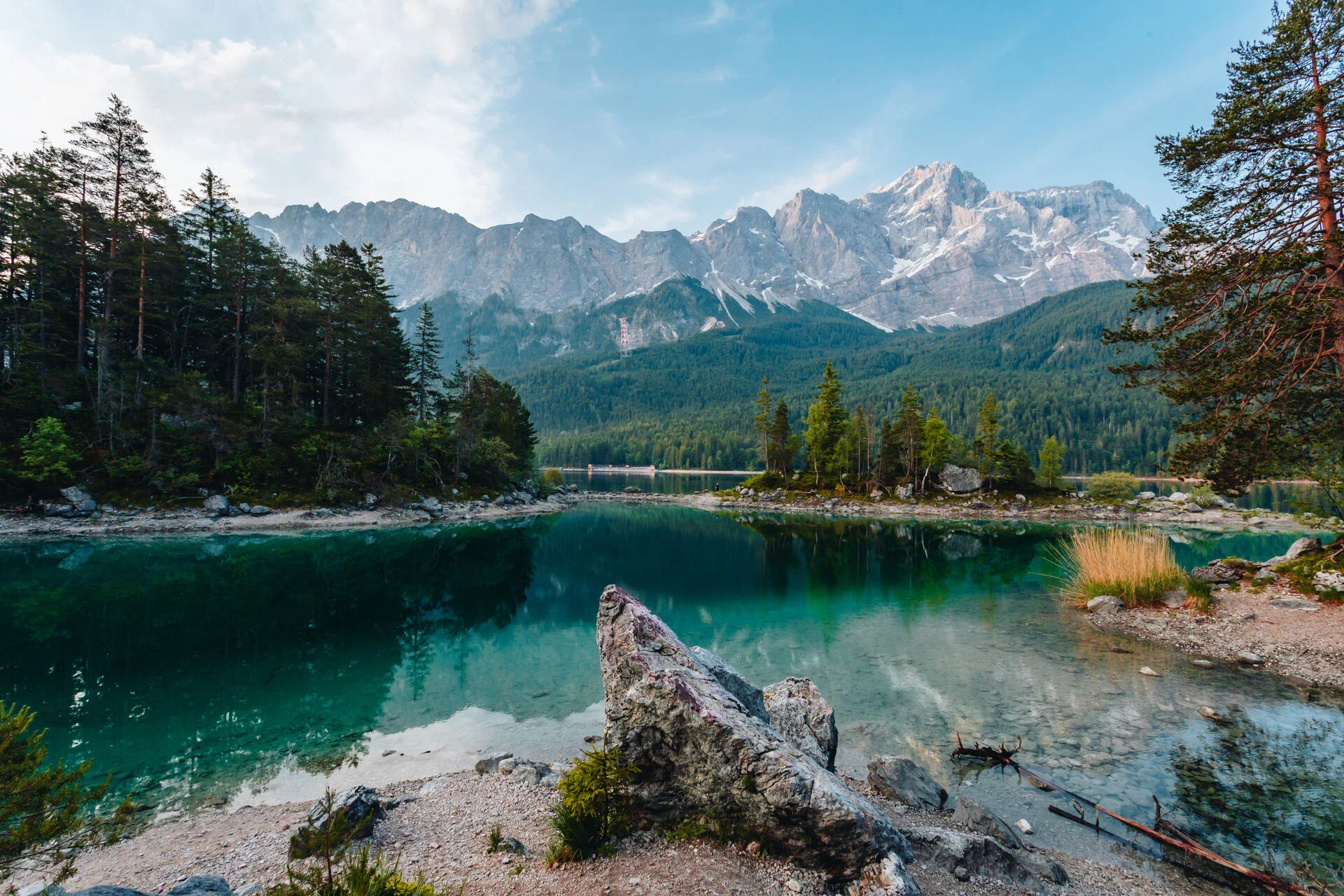Kristallklarer Bergsee umgeben von dichten Wäldern und majestätischen Bergen, die sich im ruhigen Wasser spiegeln, unter einem blauen Himmel.