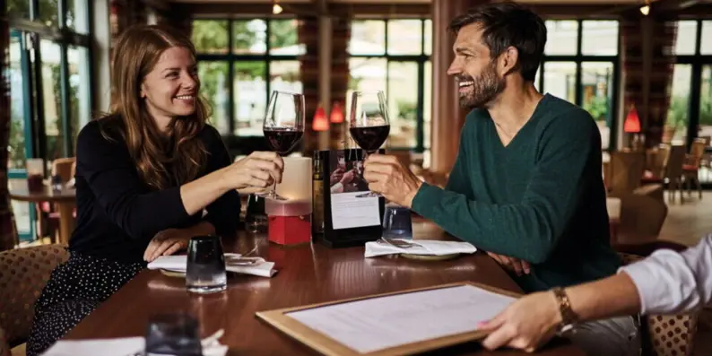Eine Frau und ein Mann sitzen in einem Bistro und stoßen mit einem Glas Rotwein an. Eine Frau und ein Mann sitzen in einem Bistro und stoßen mit einem Glas Rotwein an.