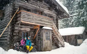Zwei Personen in Skikleidung sitzen vor einer Holzhütte im Schnee.