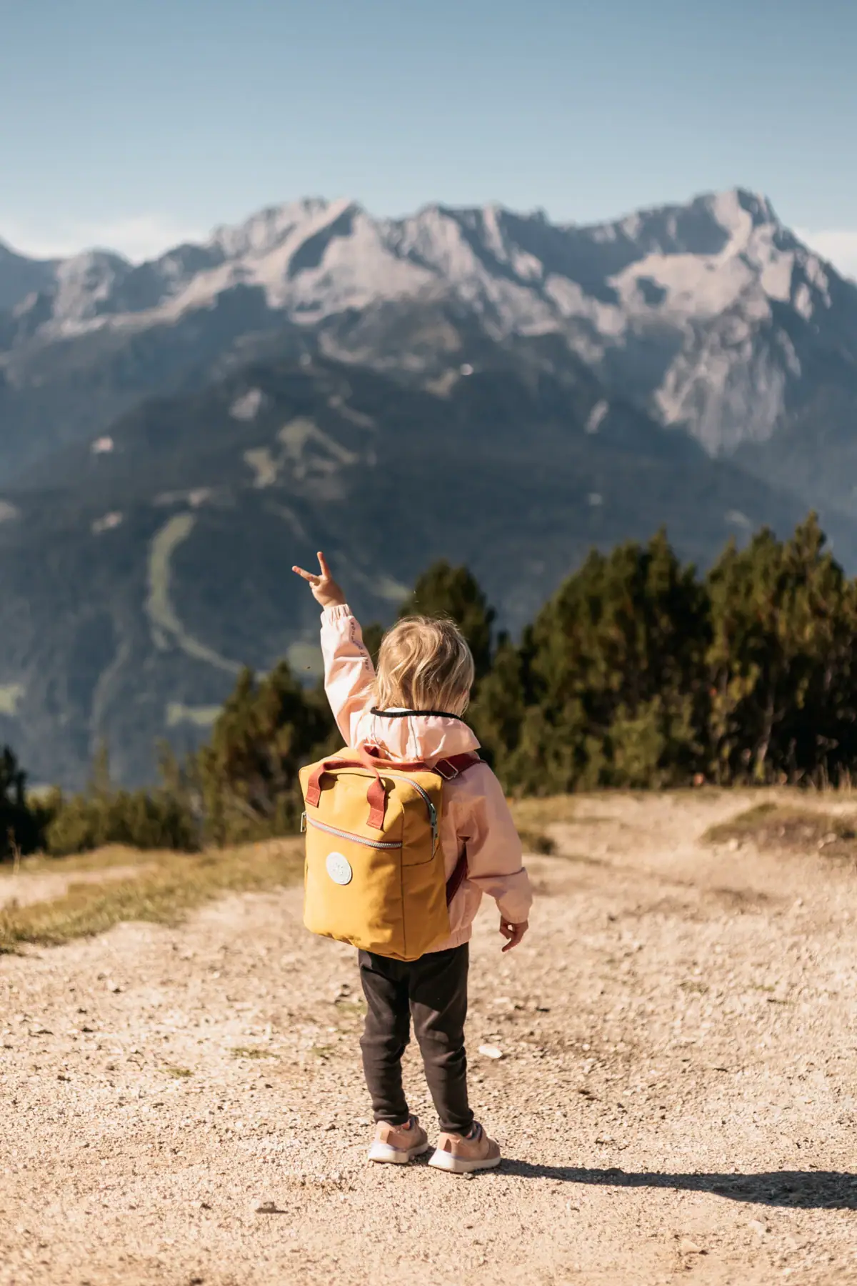 Ein Kind mit gelbem Rucksack steht auf einem Erdweg vor einer Bergkette.