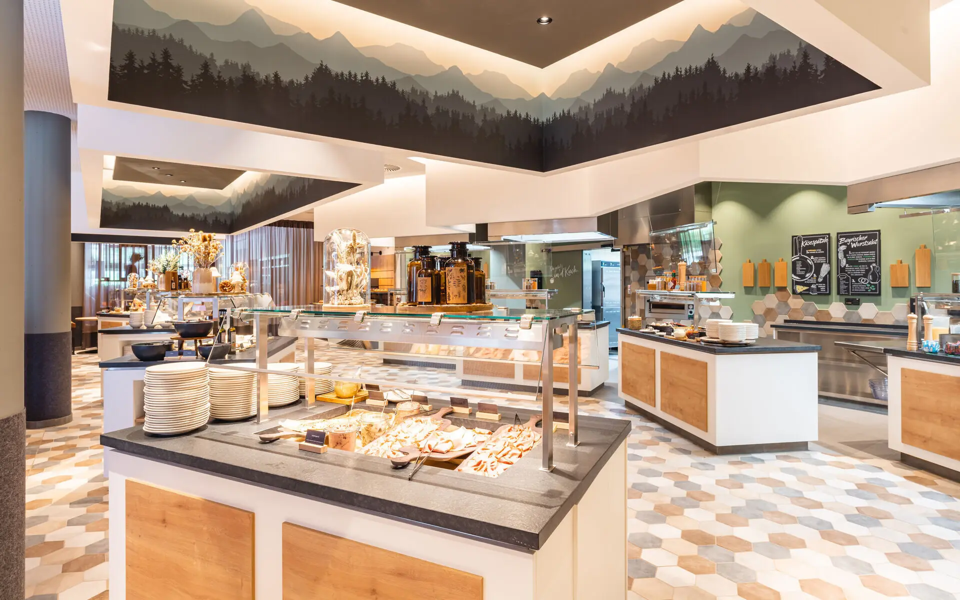 Das Bild zeigt einen modernen Buffetbereich mit Bergpanorama an der Decke, frischen Speisen und einem warmen, einladenden Design mit geometrischen Fliesen.