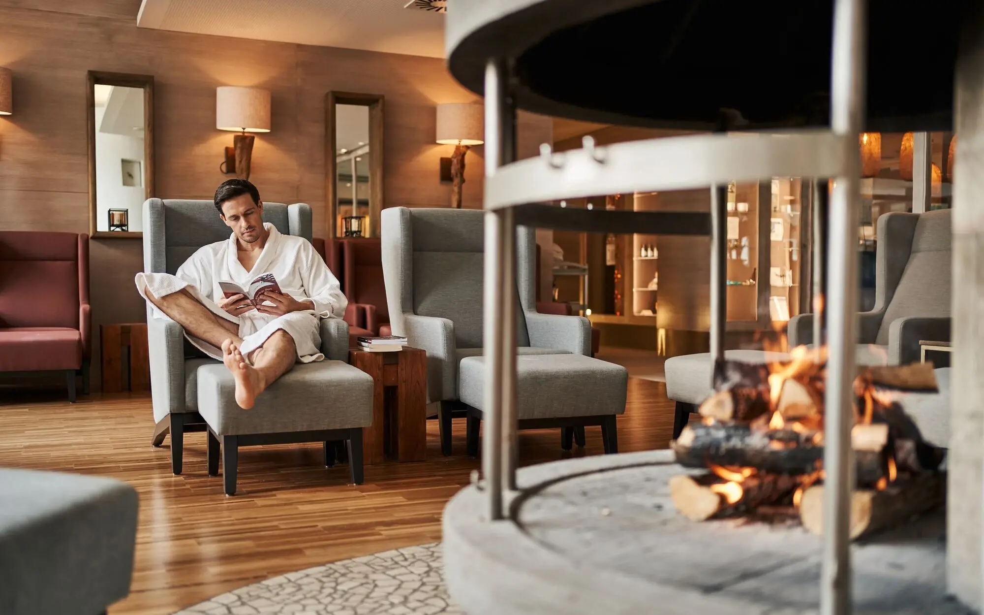 Ein Mann in einem weißen Bademantel sitzt entspannt in einem gemütlichen Sessel in einem stilvollen Lounge-Bereich und liest ein Buch. Im Vordergrund brennt ein offenes Feuer in einem modernen Kamin