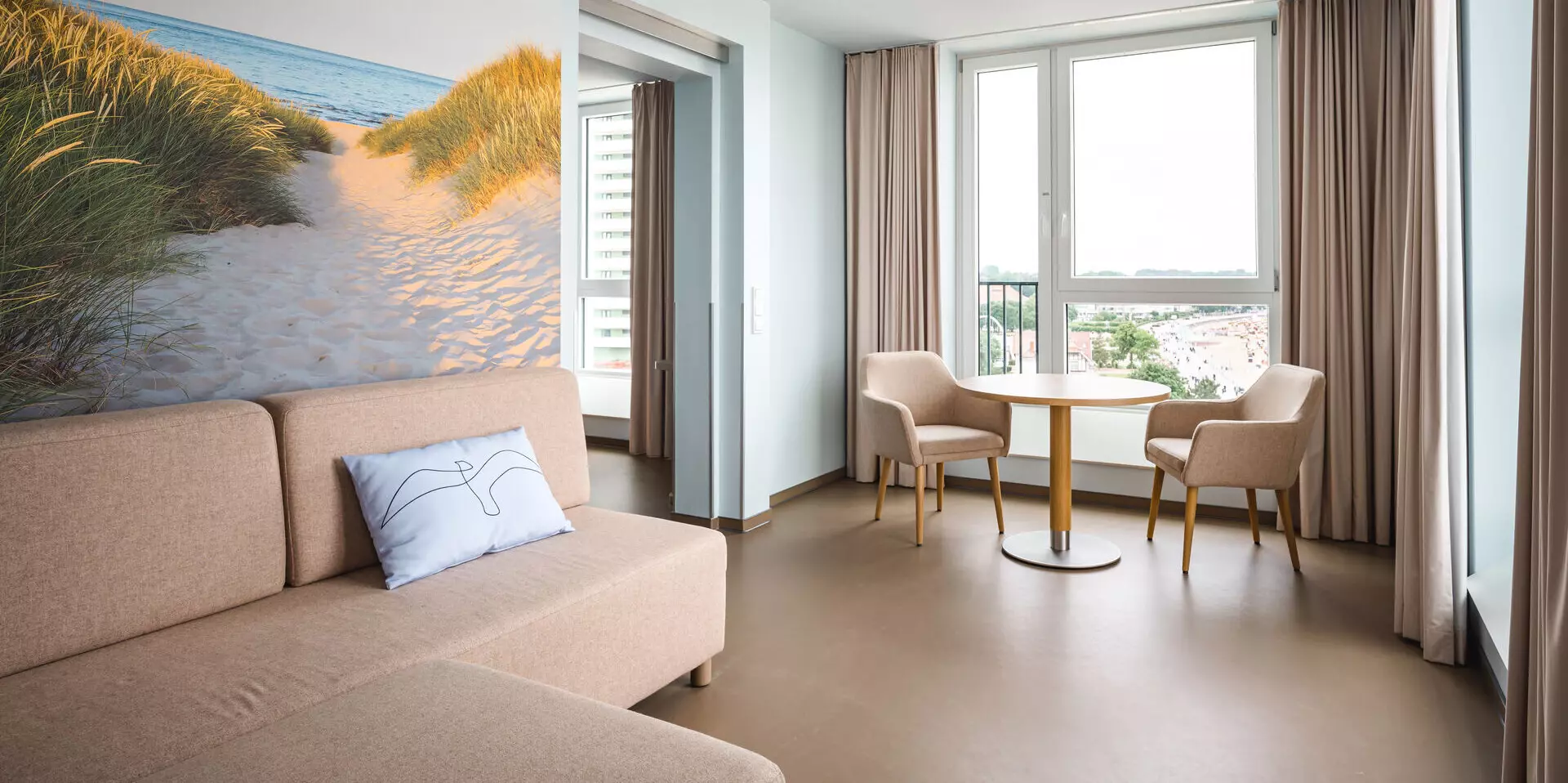 Gemütliche Hotelsuite mit einem beigefarbenen Sofa, einer hellblauen Wand mit einem Strandbild, einem kleinen runden Tisch mit zwei Stühlen sowie großen Fenstern, die einen Ausblick auf die Umgebung bieten.