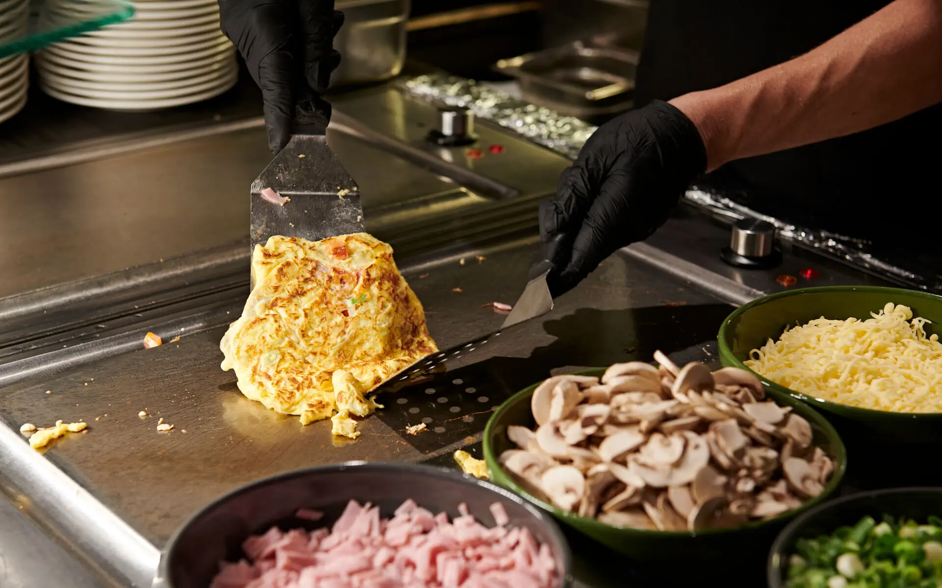 Omelette Zwei Hände mit schwarzen Handschuhen bereiten ein Omelett auf einer Kochplatte zu, um die Schälchen mit Schinken, Pilzen, Käse und weiteren Zutaten stehen.