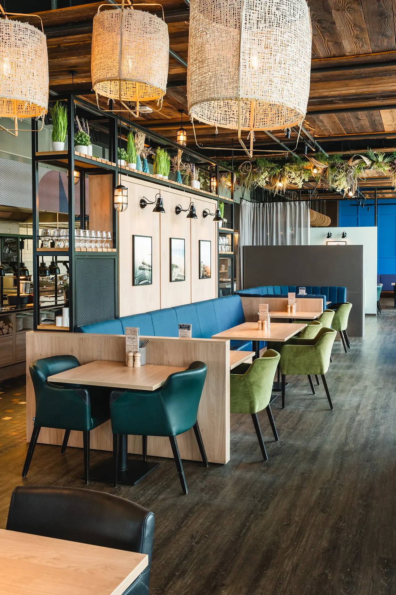Der Restaurantbereich vom aja Warnemünde mit Holzakzenten gemütlichen Sesseln in blau und grün, dunklem Boden und großen geflochtenen Lampenschirmen mit warmem, angenehmem Licht.