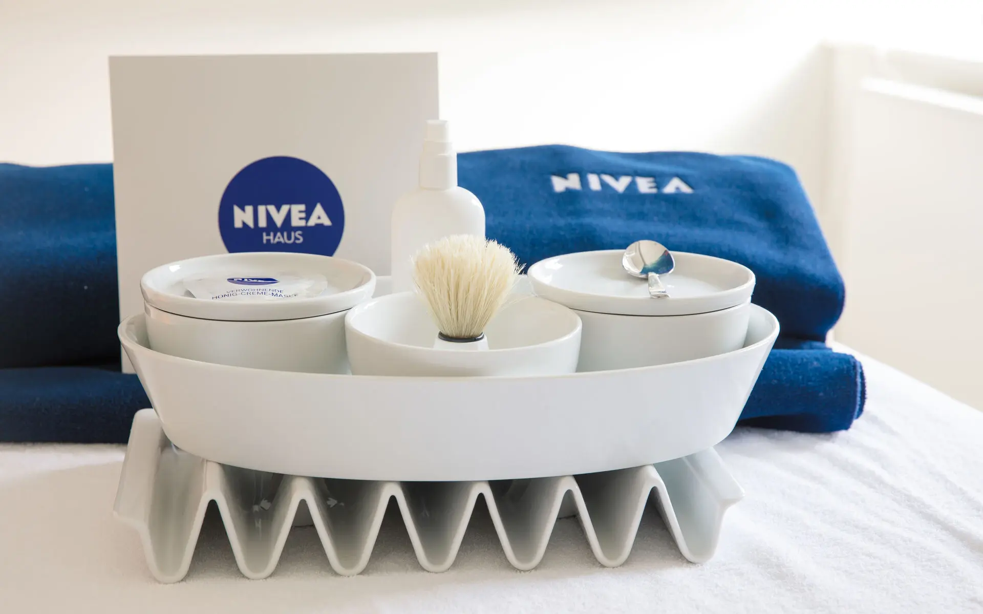 NIVEA Produkte Vor zwei dunkelblauen, gefalteten Handtüchern mit NIVEA-Logo stehen in einer weißen, ovalen Schale drei weiße, runde Schüsselchen mit Deckel. Dazwischen ist eine weiße NIVEA-Tüte mit dunkelblauem NIVEA-Logo platziert.