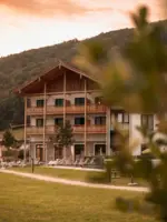 Außenansicht eines alpinen Hotels mit Holzfassade und Balkonen, umgeben von grünen Wiesen und Bäumen.Liegestühle stehen bereit und laden zum Entspannen im Freien ein, während der bewaldete Hügel im Hintergrund die natürliche Umgebung unterstreicht.