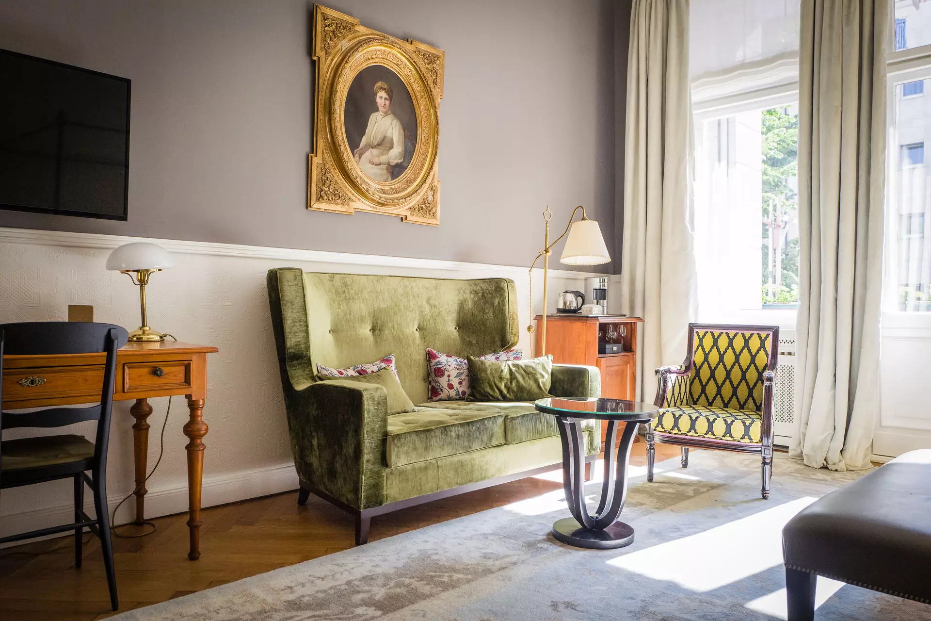 HENRI Berlin Zimmer Elegantes Hotelzimmer im HENRI Berlin mit einem samtgrünen Sofa, antikem Sessel, Holztisch, goldgerahmtem Portrait an der Wand und großen Fenstern, die den Raum mit Tageslicht durchfluten.