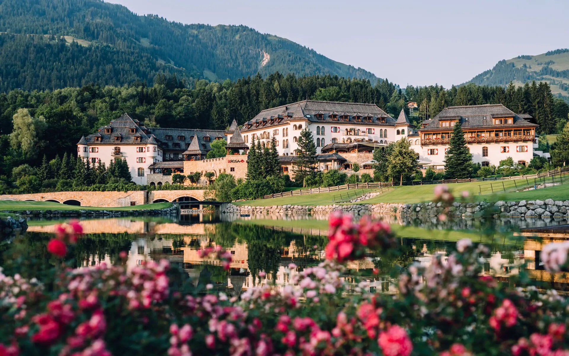A-ROSA Kitzbühel Panorama des A-ROSA Kitzbühel, umgeben von grünen Wiesen, einem idyllischen Teich mit Spiegelung und blühenden Rosen im Vordergrund, eingebettet in die malerische Berglandschaft Tirols.