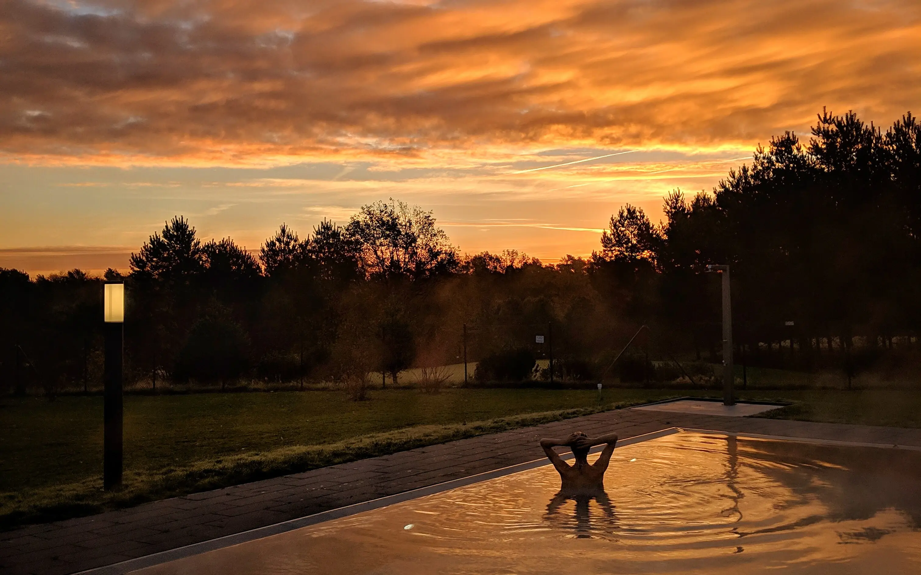 Außenpool aja Bad Saarow Eine Person schwimmt bei Sonnenuntergang in einem Pool auf dem sich die warmen Farben der tief stehenden Sonne wieder spiegeln.