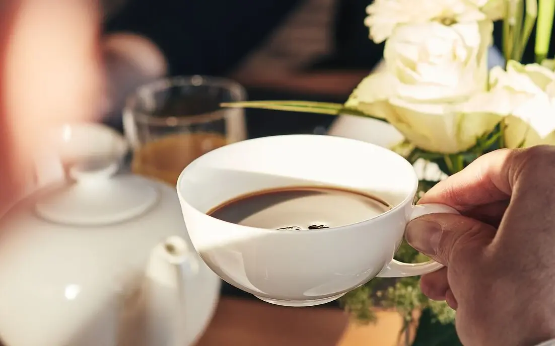 Kaffee aja Person hält eine Tasse Kaffee und im Hintergrund ist eine Kanne und Rosen zu sehen.
