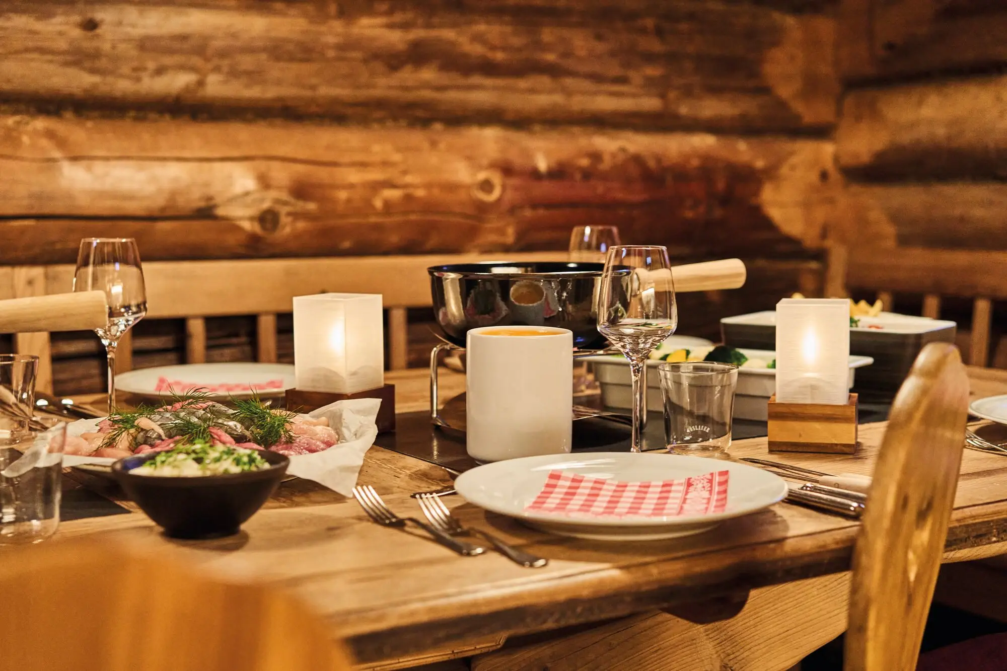 Essen im aja Werfenweng Ein rustikal gedeckter Holztisch in gemütlicher Hüttenatmosphäre, vorbereitet für ein Fondue-Essen. Auf dem Tisch stehen ein Fonduetopf, Teller mit frischen Zutaten, Gläser, Kerzenlichter und Besteck, die eine einladende und warme Stimmung erzeugen.