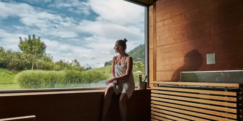 Frau sitzt in einer Sauna auf einer Fensterbank und blickt durch ein großes Panoramafenster nach draußen. Frau sitzt in einer Sauna auf einer Fensterbank und blickt durch ein großes Panoramafenster nach draußen.