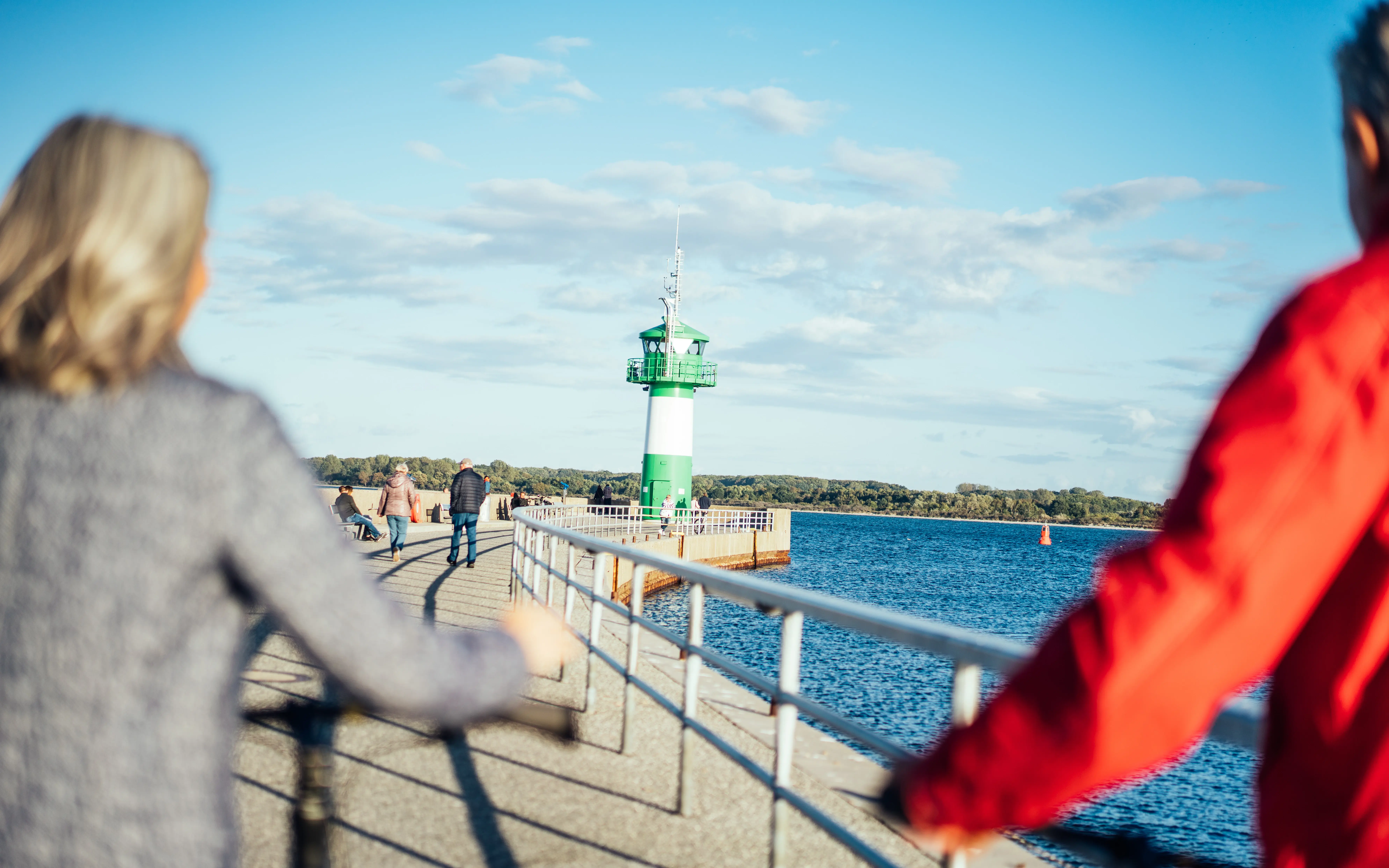Ein Paar fährt mit Fahrrädern entlang einer Mole in Richtung eines grünen Leuchtturms, bei klarem Himmel und Blick auf das Wasser.