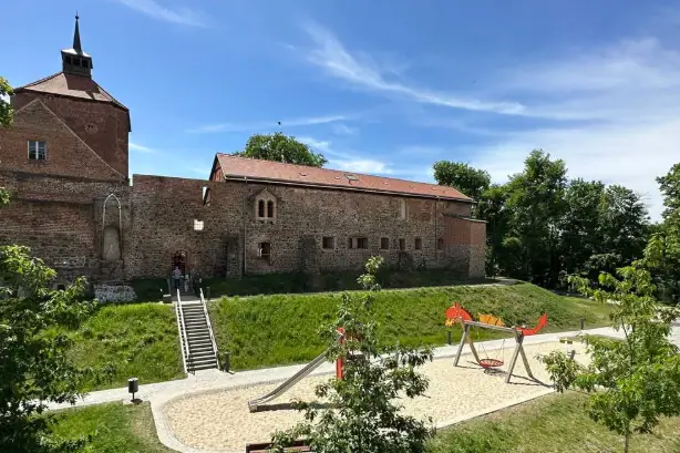 Burg Beeskow besuchen Ein Spielplatz vor einem Gebäude mit Bäumen und blauem Himmel.