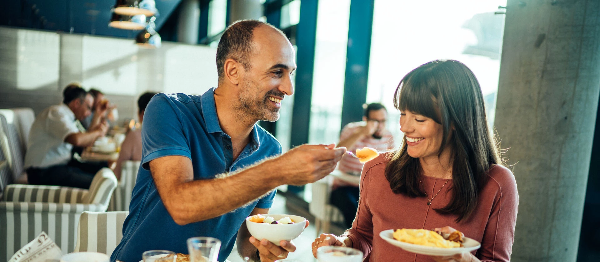 Frühstück im aja Travemünde Ein Paar sitzt an einem reichlich gedeckten Frühstückstisch mit Müsli, Früchten, Eierspeisen und Kaffeespezialitäten. Die Frau in rotem T-Shirt hält einen Teller in ihrer Hand und der Mann in blau hält ihr lachende eine Gabel entgegen.