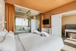 Suite im aja Warnemünde Ein Bett mit weißen Laken und Handtüchern in einem Zimmer. An der Wand der Fernseher, im Hintergrund das Bad mit Glastür