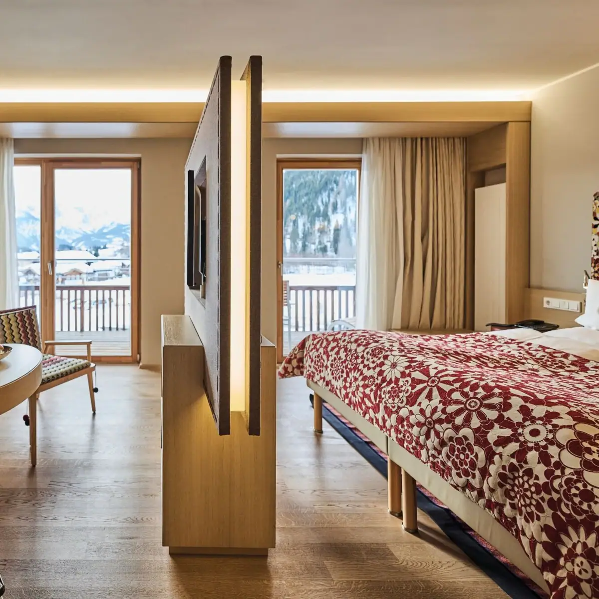 Modern eingerichtetes Hotelzimmer mit separatem Wohn- und Schlafbereich, Holzfußboden und warmen Farbtönen. Ein Sofa mit gemusterten Kissen, ein runder Holztisch mit zwei Gläsern Wein sowie ein Fernseher auf einer Trennwand dominieren den Raum.