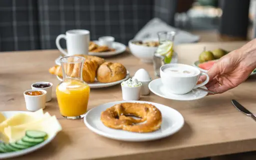 Ein gedeckter Frühstückstisch mit Brezel, Brötchen und Aufschnitt. Eine Hand stellt eine Tasse Kaffee dazu. Ein gedeckter Frühstückstisch mit Brezel, Brötchen und Aufschnitt. Eine Hand stellt eine Tasse Kaffee dazu.