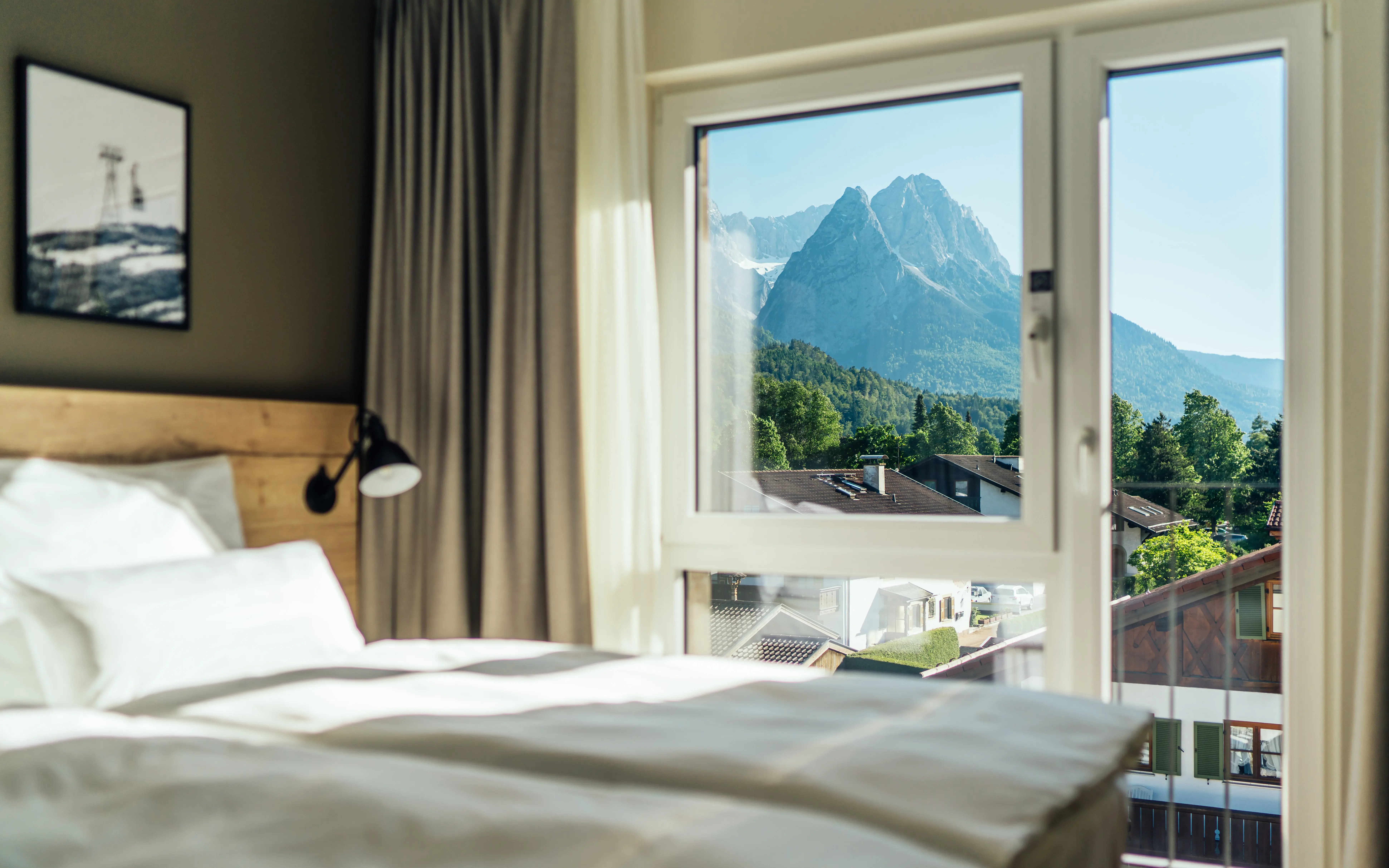 Ein Hotelzimmerausschnitt mit gemütlichem Bett neben einem großen Fenster, das den Blick auf umstehende Häuser und die dahinter liegende Natur frei gibt.