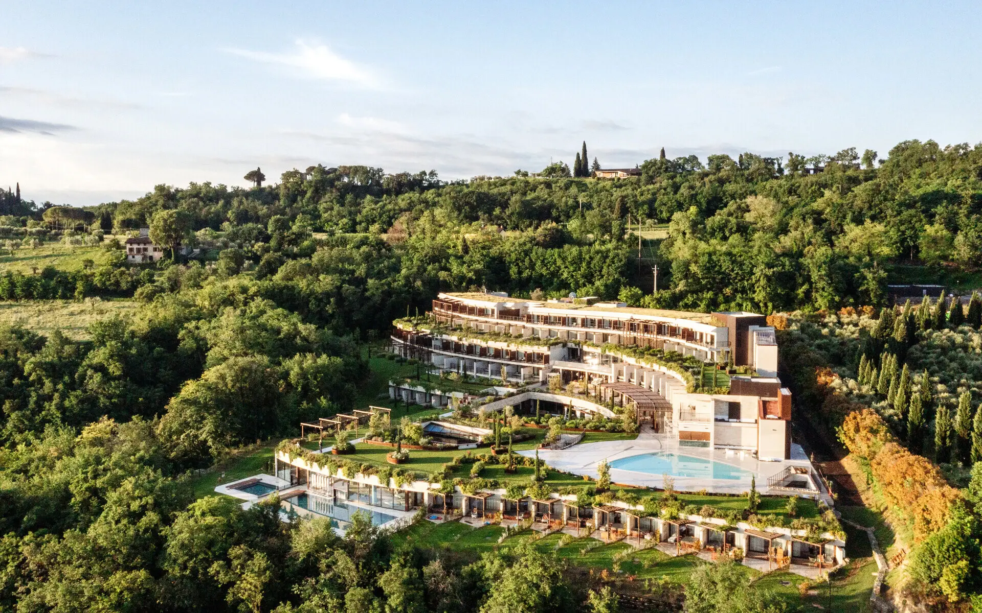 A-ROSA Gardasee Vogelperspektive auf das A-ROSA Gardasee, harmonisch eingebettet in die grüne Hügellandschaft oberhalb des Gardasees, mit terrassenförmigen Gebäuden, einem großzügigen Poolbereich und mediterraner Vegetation.