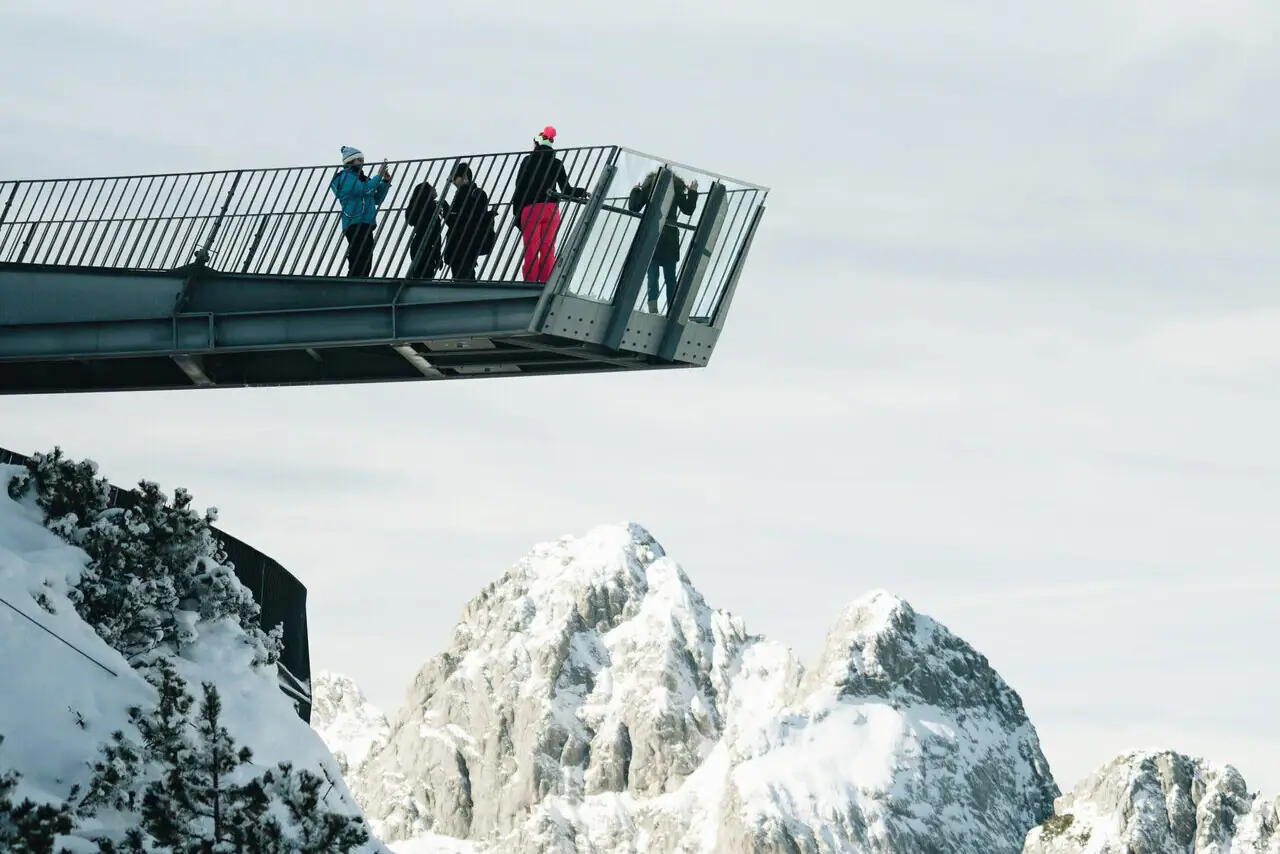 AlpspiX Menschen stehen auf einer Brücke über einem verschneiten Berg.