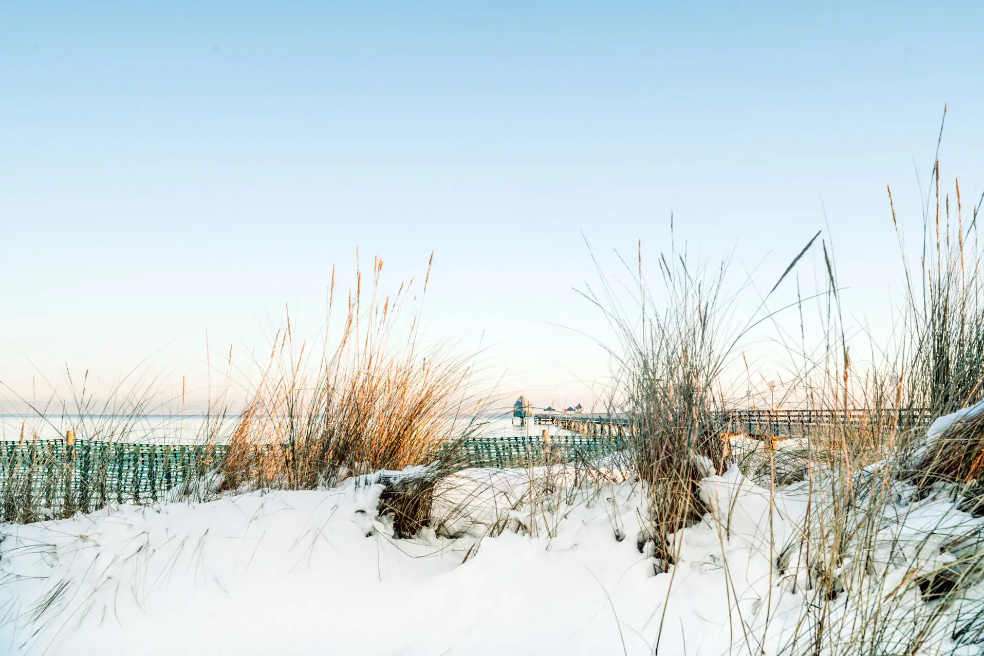 Grömitz im Schnee Schneebedeckter Boden mit hohem Gras und einem Pier im Hintergrund.
