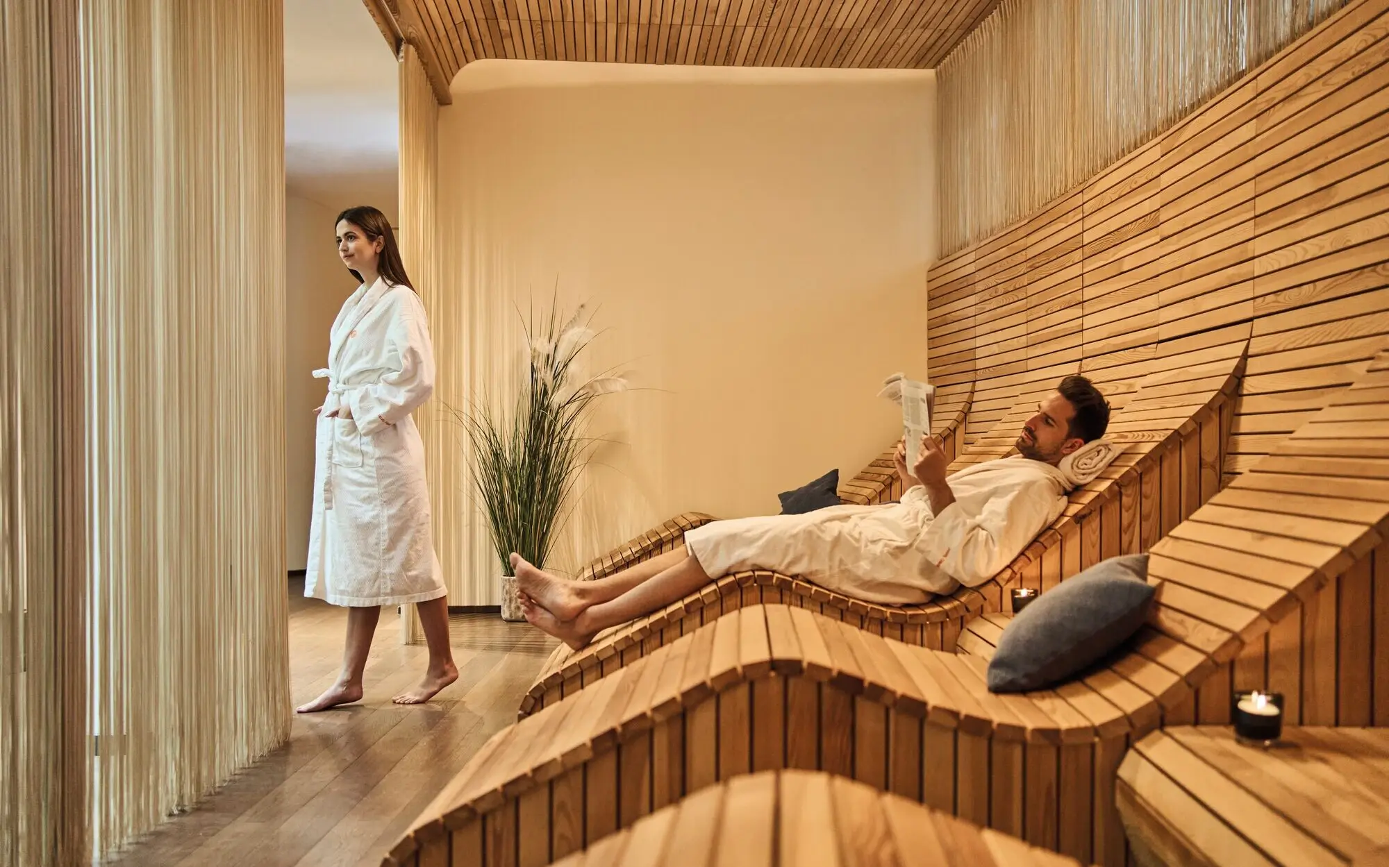Wellness aja Werfenweng Ein entspannter Spa-Bereich mit geschwungenen Holzliegen und einem Paar in weißen Bademänteln. Der Mann liegt gemütlich auf einer Liege und liest, während die Frau ruhig am Fenster steht.