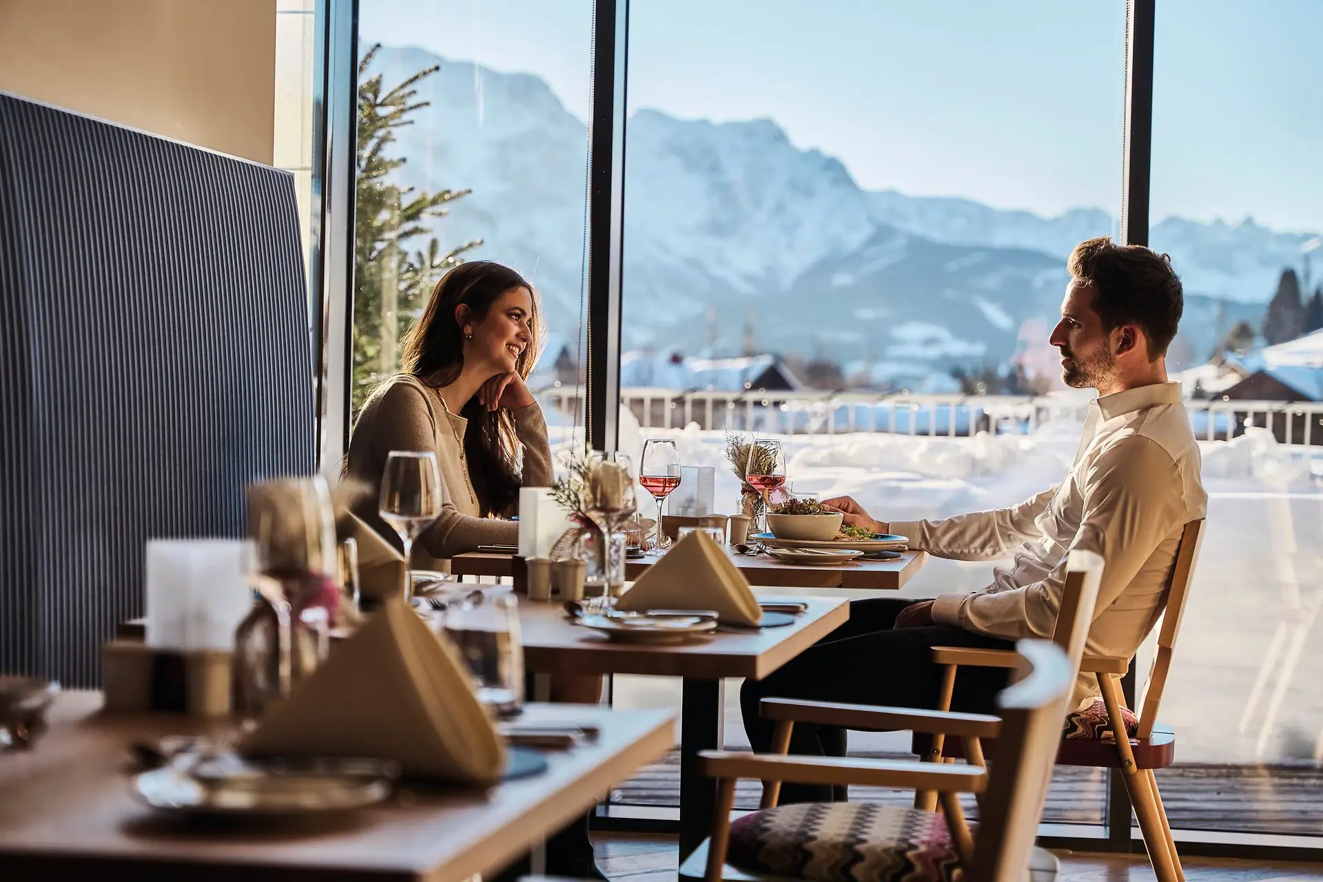 Feinspitz Restaurant Ein Mann und eine Frau sitzen an einem Tisch mit Essen und Weingläsern, im Hintergrund sind de Berge zu sehen.