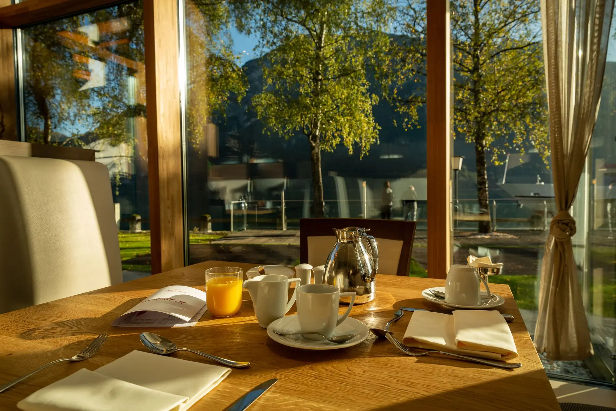 Frühstück im Fürstenhaus am Achensee Ein Tisch mit einer Glaskanne, Teetassen und Untertassen an einem Tisch neben einem Fenster mit Blick auf Bäume.