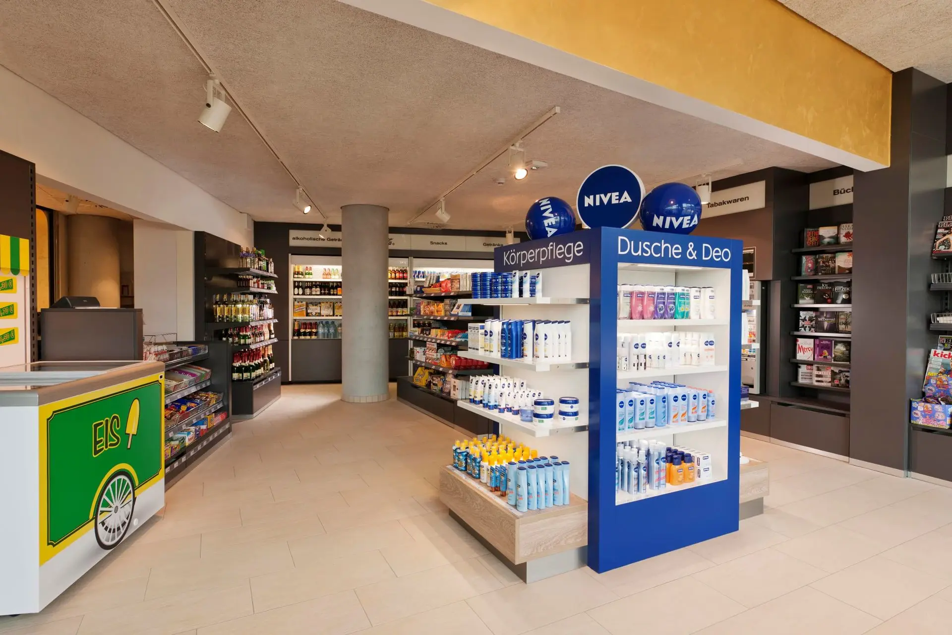 Ein kleiner Laden mit dunkelgrauen Wänden und hellem Boden. Im hinteren Bereich befinden sich Kühlregale und in der Mitte des Raumes steht ein blauer Niveaaufsteller mit Produkten. Kleine Lampen an der Decke spenden warmes Licht.