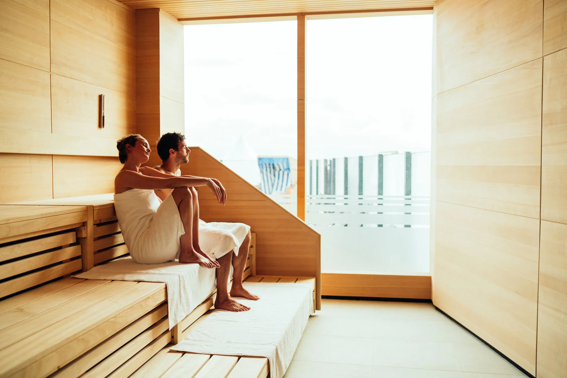 Ein Mann und eine Frau sitzen in weiße Handtücher gewickelt in einer Holzsauna und entspannen. Das Licht ist warm und durch ein Fenster fällt Tageslicht herein.
