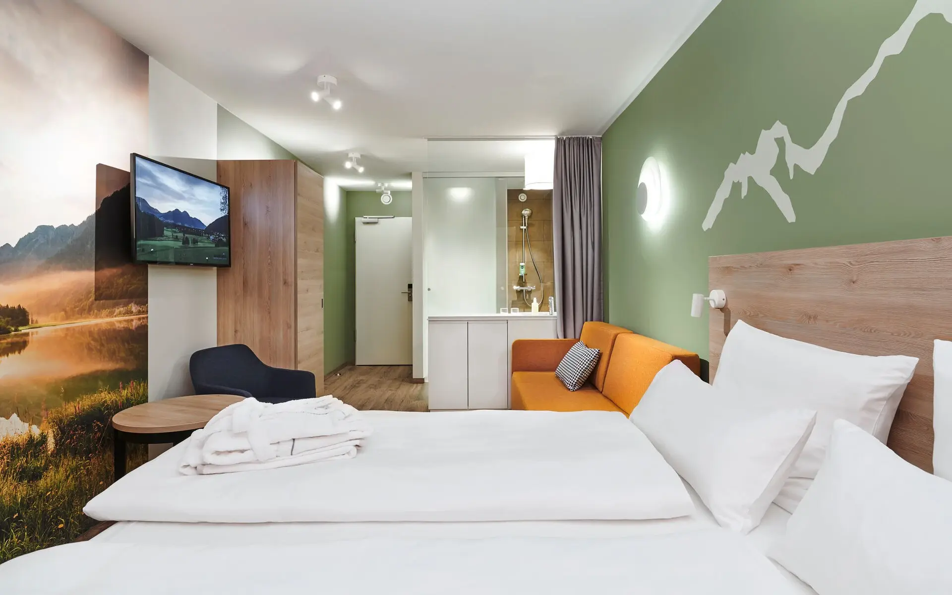 Ein gemütliches Hotelzimmer mit einem Bett vor einer hell grünen Wand mit aufgezeichneter Bergkette, einem senffarbenen Sofa und einem Glasbadezimmer im Hintergrund.