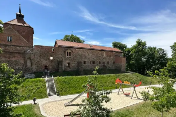 Burg Beeskow besuchen Ein Spielplatz vor einem Gebäude mit Bäumen und blauem Himmel.