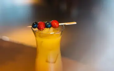 Auf einem Holztisch steht ein hohes Cocktailsglas mit einem orangenen Cocktail. Auf dem Rand stützt ein Holzstäbchen mit aufgefädelten Beeren.