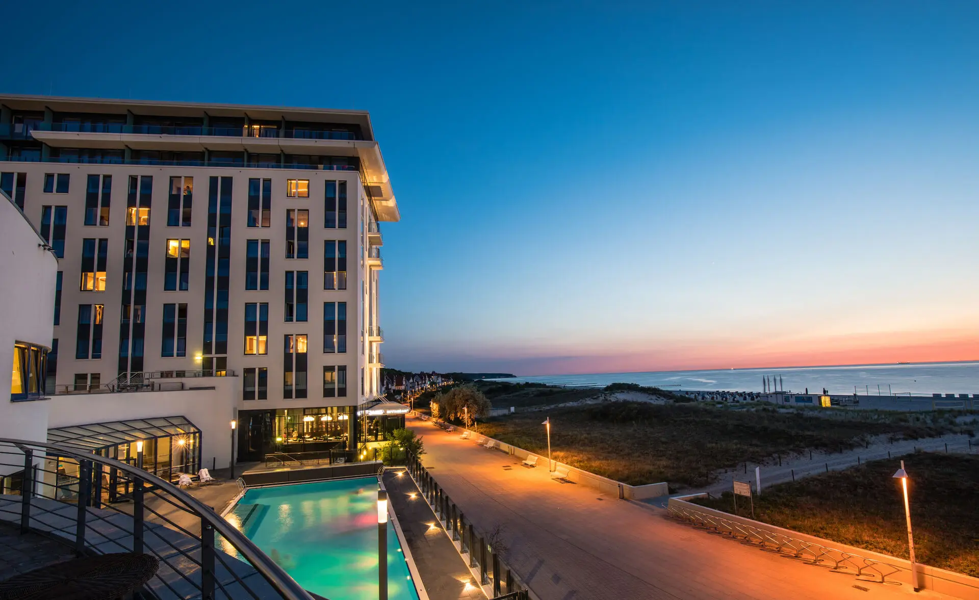 Das aja Warnemünde, ein weißes Hotel mit vielen Fenstern und befeuchtetem Außenpool bei untergehender Sonne, die das angrenzende Meer und den Himmel in warme Farben taucht.