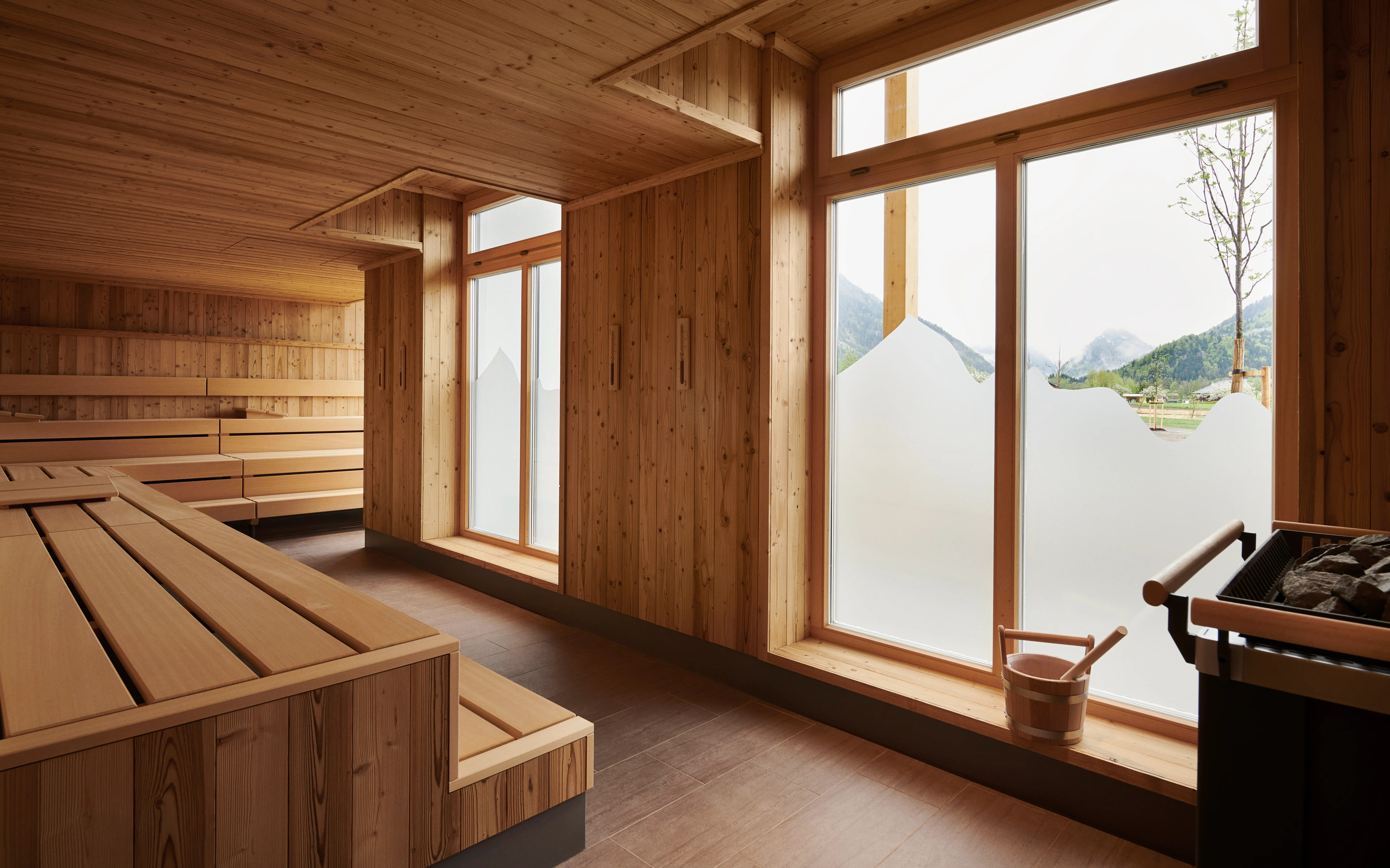 Saunabereich im aja Ruhpolding Helle Sauna mit Holzverkleidung und großzügigen Fenstern, die einen leicht getrübten Blick auf die umliegenden Berge ermöglichen.