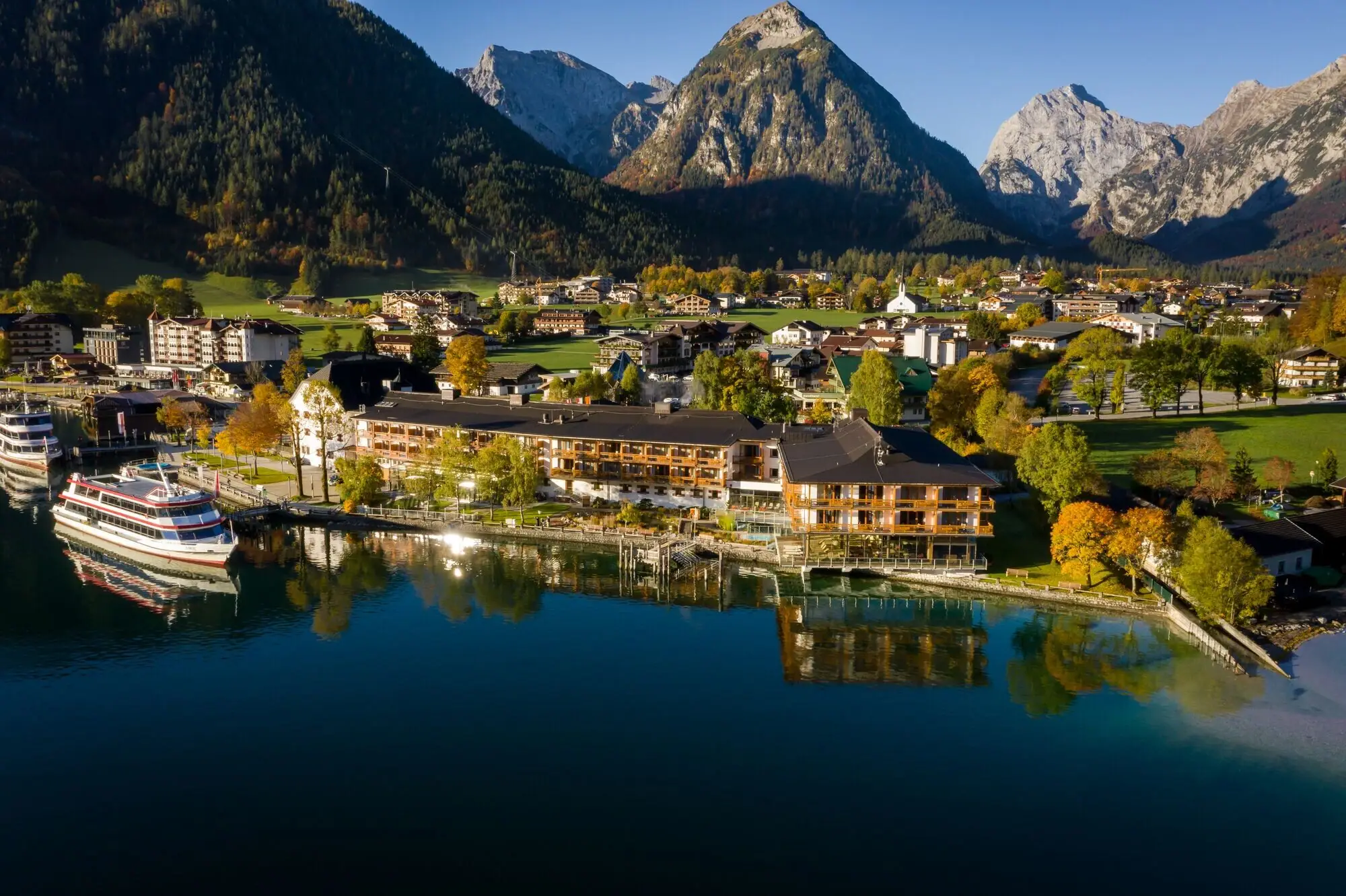 aja Achensee Hotel vor Bergpanorama Eine Stadt am Wasser mit Bäumen und einem Boot im Vordergrund.